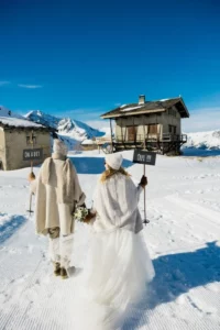 Les futurs mariés habillés en vêtements d"hiver blancs se dirigeant vers deux chalets sur la montagne enneigée